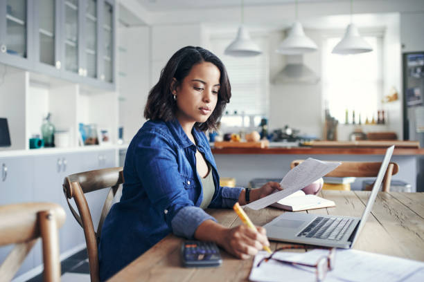 Shot of a young woman using a laptop and going through paperwork while working from home